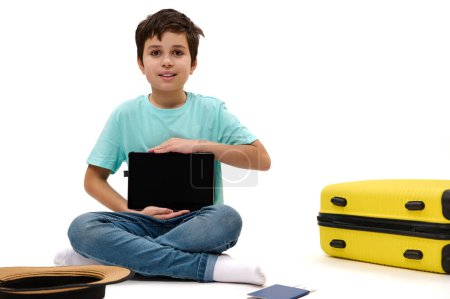 Foto de Niño viajero adolescente en camiseta azul, muestra ante la cámara una tableta digital con pantalla en blanco con espacio para insertar su texto publicitario o aplicaciones móviles, sentado cerca de la maleta amarilla sobre fondo blanco - Imagen libre de derechos