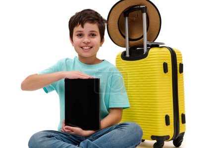Foto de Handsome teenage boy, young tourist traveler, holding digital tablet with empty space - mockup - for advertising text, smiling looking at camera, sitting near his yellow suitcase over white background - Imagen libre de derechos