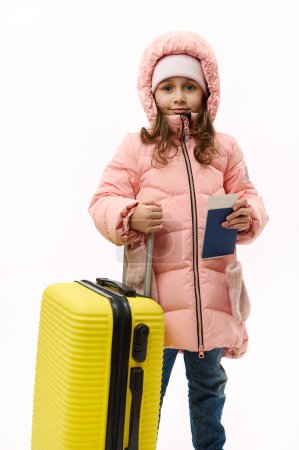 Téléchargez les photos : Full-length portrait: Beautiful little girl in pink down coat, going for vacations, looking at camera, posing with suitcase and boarding pass on white background with copy space. Travel Trip Journey - en image libre de droit