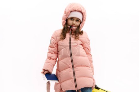 Téléchargez les photos : Adorable European child girl in pink down coat, looks at camera, poses with suitcase and boarding pass over white background. Kids going for holidays. air flight. Travel Trip Journey. Winter holidays - en image libre de droit
