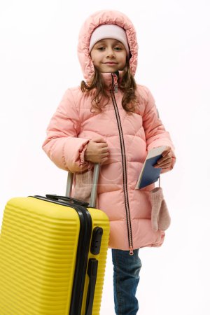 Téléchargez les photos : Lovely baby girl in pink warm down coat and blue denim jeans, going on vacations smiles cutely looking at camera, holding boarding pass and flight ticket, posing with yellow suitcase, white background - en image libre de droit