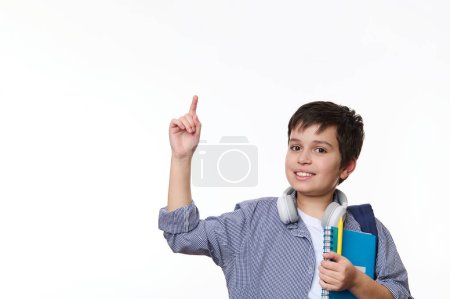 Nahöstlicher Teenager in Freizeitkleidung, der positive Emotionen ausdrückt, auf kopierte Werbeflächen zeigt, in die Kamera lächelt, mit Schulmaterial und Rucksack vor weißem Hintergrund posiert
