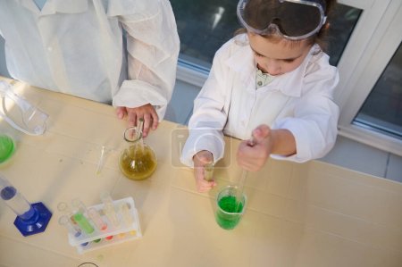 View from above of a smart school girl in white lab coat, using graduated pipette, dripping few reagents into a beaker with green chemical liquid solution, doing experiments in chemistry laboratory