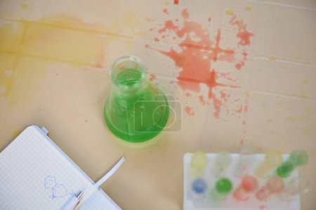 Téléchargez les photos : Flat-bottomed laboratory flask with green liquid, a tripod with test tubes with colorful chemicals and reagents and solutions spilled on a table in a chemical laboratory. Top view. Chemistry class - en image libre de droit