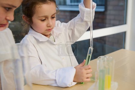 Téléchargez les photos : Smart Caucasian 5-6 years old little child , schoolgirl wearing white labo gown, dripping few reagent into a test tube with chemical liquid solution w, using a graduated pipette in a school laboratory - en image libre de droit