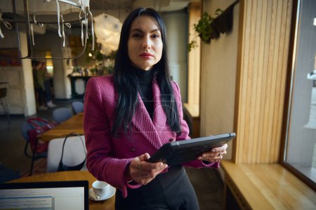 Téléchargez les photos : Pleasant charming Caucasian woman, executive manager, restaurateur, dressed in stylish formal wear, holding digital gadget in hands, looking at camera and welcoming clients on newly opened restaurant - en image libre de droit