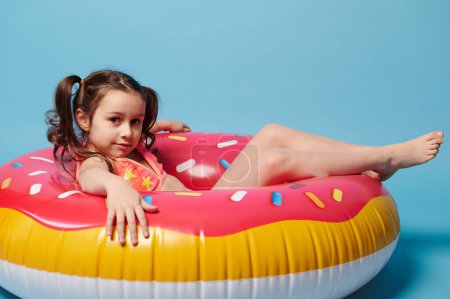 Photo for Lovely little child girl with two ponytails, wearing a stylish swimsuit, looking at camera while lying on an inflatable donut swimming ring, a colorful lifebuoy, isolated over blue color background. - Royalty Free Image