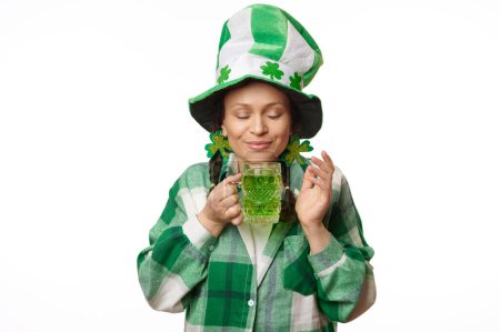 Atractiva mujer en sombrero de carnaval con hojas de trébol verde, disfrutando del sabor de la cerveza verde mientras celebra el Día de San Patricio en un pub posando con los ojos cerrados sobre un fondo blanco aislado