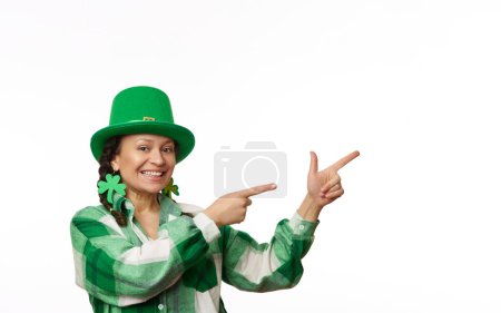 Cheerful positive dark-haired woman in green carnival clothes for St. Patricks pub party, pointing her index finger at copy space for ads on white background. March 17. Irish culture and traditions