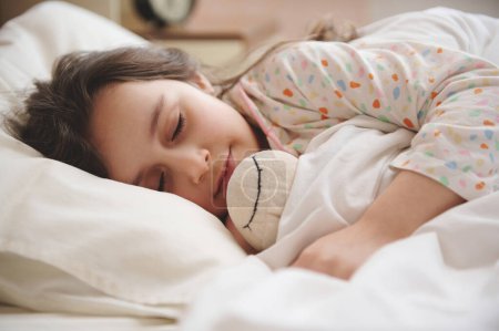 Retrato de cerca de una niña caucásica de 5 años encantadora en pijama con puntos de colores, abrazando suavemente a su felpa oveja de juguete, mientras duerme en la cama en el interior de un dormitorio luminoso. Infancia