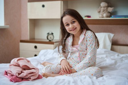 Téléchargez les photos : Portrait d'enfant d'âge élémentaire caucasien, heureuse petite fille en pyjama blanc à pois colorés, assise sur le lit dans sa chambre, près de ses vêtements pliés, souriant en regardant la caméra - en image libre de droit