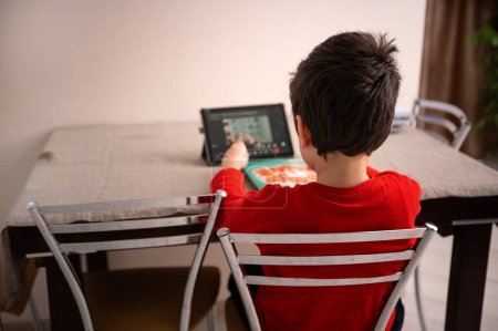 Rear view of a Caucasian 10 years child, schoolboy doing homework, sitting at table and watching online lesson on a digital tablet. Children. Homeschooling. Distance education and e-learning concept