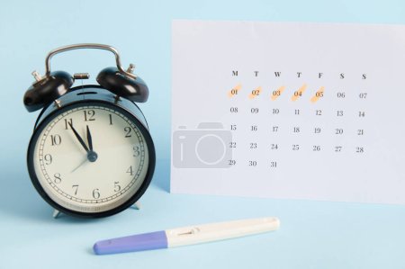 Positiver Inkjet-Schwangerschaftstest, schwarzer Retro-Wecker und weißer Kalender mit Daten der letzten Menstruation auf blauem Hintergrund. Frauengesundheit, Eisprung, Fruchtbarkeit, Schwangerschaftsplanung