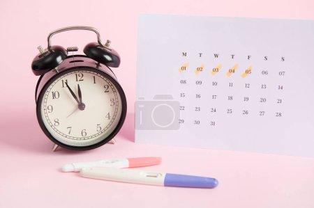 Bodegón con kits de prueba de embarazo positivos, despertador negro y calendario blanco con las fechas de la última menstruación marcadas, aisladas sobre fondo pastel rosa. Salud y ovulación de las mujeres
