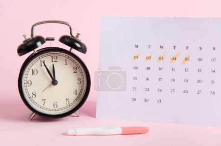 Stillleben mit schwarzem Vintage-Wecker, Kalender mit Tagen der letzten Menstruation und positivem Inkjet-Schwangerschaftstest auf rosa Pastell-Hintergrund. Schnelle Diagnose der Schwangerschaft zu Hause