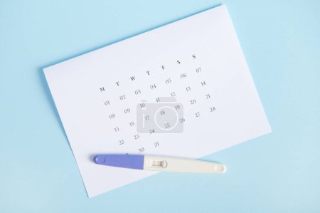 Photo for Inkjet pregnancy test showing two stripes on white clean calendar isolated on pastel blue background. Calculation of ovulation day. Planning maternity. Gynecology and womens health concept. Top view - Royalty Free Image