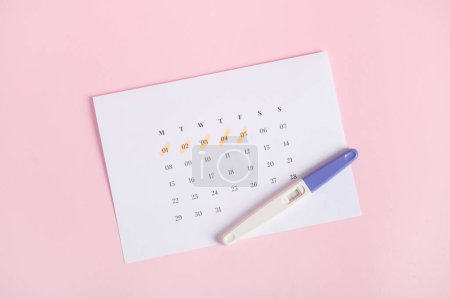 Photo for Pregnancy test showing two stripes on calendar with the dates of the last menstruation marked, pink background. Calculation of ovulation day. Planning maternity. Gynecology and womens health concept - Royalty Free Image
