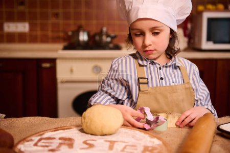 Photo for Caucasian 5-6 years old adorable little toddler girl, in white chefs hat and beige kitchen apron, standing at a table with a dough on a floured wooden board with words Easter. Child learns cooking - Royalty Free Image
