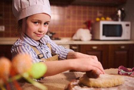 Photo for Beautiful little girl 5 years old, wearing white chefs hat and beige apron, smiling cutely looking at camera, standing at kitchen table and rollling out the dough. Blurred Easter eggs on foreground - Royalty Free Image