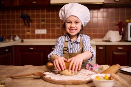 Photo for Caucasian mischievous little child girl using rolling pin, rolls out dough on a floured wooden board, preparing homemade gingerbread cookies for Easter holiday, smiling broadly looking at camera - Royalty Free Image