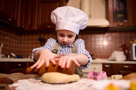 Photo for Caucasian lovely little toddler girl in white chefs hat, rolls out dough on floured wooden board, using rolling pin, preparing delicious gingerbread cookies for Easter holidays in the home kitchen - Royalty Free Image