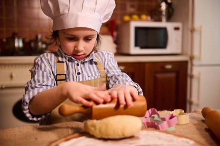 Photo for Adorable Caucasian 5-6 years child girl, little chef confectioner in cook uniform, standing at kitchen island and using rollin pin, rolls out dough on floured wooden board, prepares Easter gingerbread - Royalty Free Image