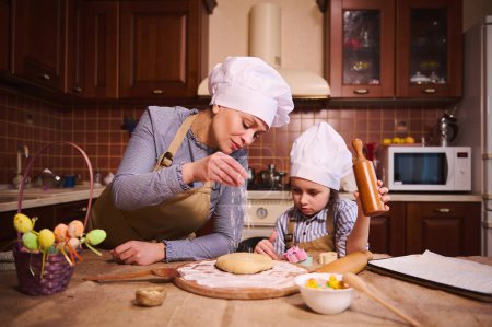 Photo for Multi-ethnic pregnant pretty woman, loving mom in chefs hat and beige apron, standing with her lovely daughter at kitchen island, flouring dough, preparing cakes and panettone for Easter holiday - Royalty Free Image