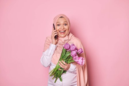 Photo for Beautiful Muslim pregnant woman in hijab, holding bouquet of purple tulips, talking on mobile phone, expressing amazement, looking at camera, isolated on pink background. Happy Mothers Day concept - Royalty Free Image