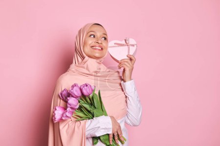 Téléchargez les photos : Heureuse femme musulmane arabe portant le hijab rose, tenant un beau bouquet de tulipes violettes et une boîte cadeau en forme de coeur, souriant regarder cutly côté, isolé sur fond rose avec espace publicitaire copie - en image libre de droit