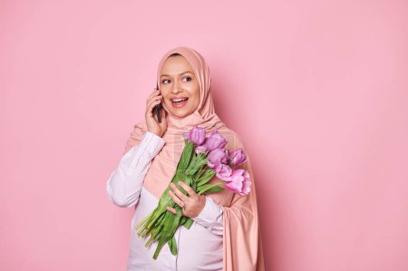 Photo for Gorgeous happy muslim young pregnant woman, dressed in elegant pink hijab, holding bouquet of tulips and talking on smartphone, smiling looking away isolated on pink. Pregnancy and maternity concept - Royalty Free Image