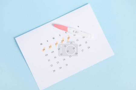Photo for Rapid diagnosis of pregnancy at home. Top view of inkjet pregnancy test with two bars on white calendar with last menstruation dates marked, isolated on blue background. Womens heath concept - Royalty Free Image