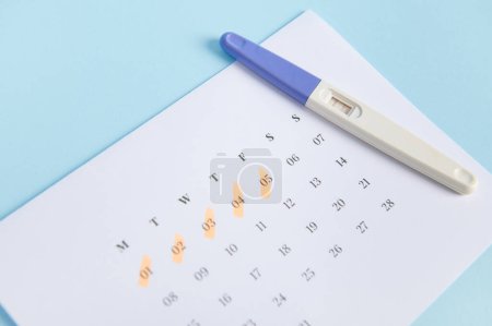 Photo for The concept of calculating ovulation and fertile days, planning maternity and diagnosis pregnancy. Inkjet pregnancy test kit on a white calendar with dates of last menstruation marked, blue background - Royalty Free Image
