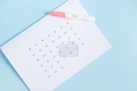 Photo for The concept of calculating ovulation and fertile days, planning maternity and diagnosis pregnancy. Inkjet pregnancy test kit on a white calendar with dates of last menstruation marked, pink background - Royalty Free Image
