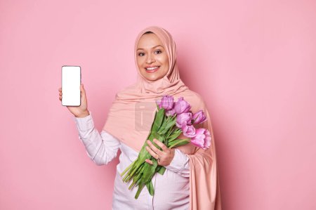 Photo for Smiling pregnant Muslim woman in pink hijab, holds a bouquet of tulips, shows at camera a smartphone with white blank screen with copy space for insert your advertisement, isolated on pink background - Royalty Free Image