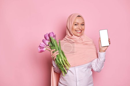 Photo for Gorgeous Middle-Eastern Muslim woman in pink hijab, showing at camera a smartphone with white blank digital screen with ad space for mobile application, posing with tulips on isolated pink background - Royalty Free Image