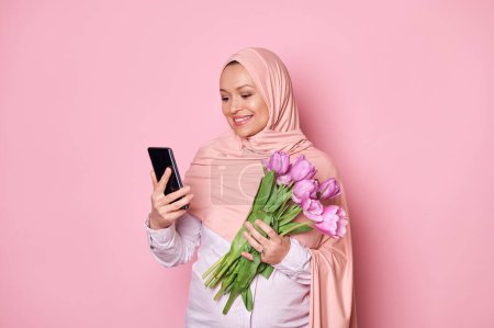 Photo for Arab Muslim pregnant woman in pink hijab, holding a bouquet of purple tulips and checking her smartphone, downloading new mobile application, text messaging, smiling on isolated pink color background - Royalty Free Image