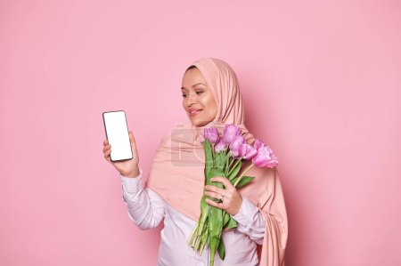 Photo for Happy pregnant Arab Muslim woman in pink hijab, with a bouquet of tulips, looking at smartphone with white blank screen, with free advertising space for mobile application, isolated on pink background - Royalty Free Image