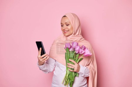 Photo for Middle-Eastern Muslim pregnant woman in pink hijab, using smartphone, scrolling news feed, checking social media content, testing new mobile app, posing with a bouquet of tulips, isolated background - Royalty Free Image