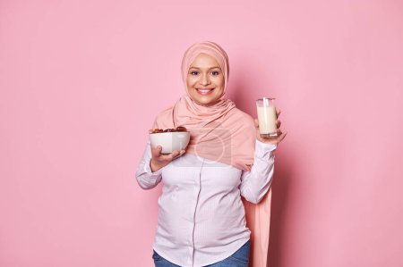 Téléchargez les photos : Grossesse et calcium. Joyeux Arabe musulman enceinte en hijab rose, tenant un verre de lait végétalien et un bol avec des dates douces fraîches, souriant à la caméra, isolé sur fond de couleur rose. Espace libre - en image libre de droit