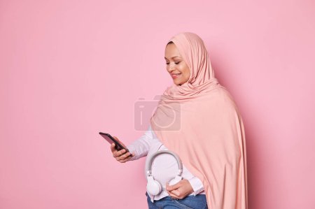 Photo for Pretty Muslim woman in pink hijab, scrolling play list on smartphone and putting headphones on her tummy, enjoying happy pregnancy over pink color background. Copy ad space - Royalty Free Image