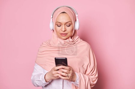 Photo for Close-up portrait of a Middle-Eastern Muslim woman in pink hijab, using mobile phone, scrolling playlist, checking mobile apps, enjoying listening to music in headphones, isolated over pink background - Royalty Free Image