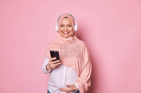 Photo for Happy pregnant Arabian Muslim woman in pink hijab and headphones, checking new mobile application on smartphone, rejoicing at feeling first baby kicks, smiling looking at camera over pink background - Royalty Free Image