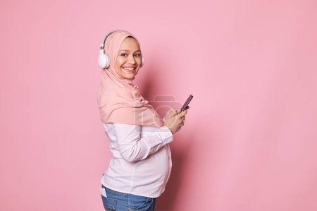 Photo for Young happy pregnant Muslim woman in pink hijab and headphones, checking new mobile application on her smartphone, smiling with beautiful toothy smile looking at camera, isolated on pink background - Royalty Free Image