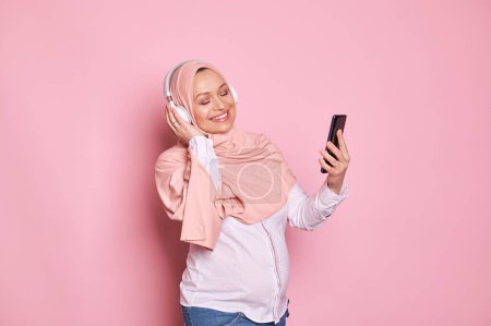 Photo for Beautiful delightful pregnant Muslim woman in pink hijab and headphones, enjoys a soothing music, smiling posing with a smartphone, scrolling playlist with cool tracks, isolated on pink background - Royalty Free Image