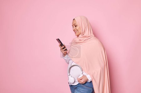 Photo for Happy pregnant Muslim woman in pink hijab, puts headphones on belly, uses smartphone and takes notes in new mobile apps, records babys movements, smiles isolated on pink background. Happy pregnancy - Royalty Free Image