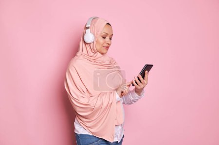 Photo for Pregnant Muslim woman in pink hijab, checking mobile application on smartphone, scrolling playlist and listening to music on wireless headphones, isolated over pink background. Copy space - Royalty Free Image