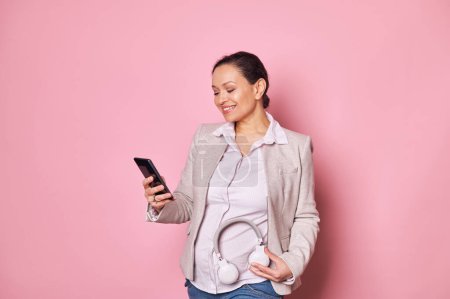 Photo for Delightful pregnant pretty woman puts headphones on belly, uses smartphone and takes notes in new mobile apps, records babys movements, smiles isolated on pink background. Happy pregnancy - Royalty Free Image