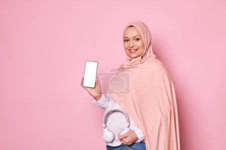 Photo for Muslim pregnant woman in pink hijab, sharing soothing music with her upcoming new baby, putting headphones on her belly, smiling, showing a smartphone with white blank screen with space for mobile app - Royalty Free Image