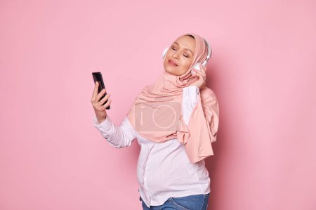 Photo for Delightful young Muslim pregnant woman in pink hijab, enjoys listening to soothing music on headphones, posing with her eyes closed and a mobile phone in hands, isolated over pink background - Royalty Free Image