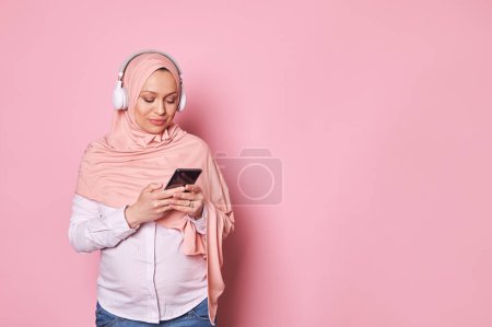 Photo for Middle-eastern beautiful Muslim pregnant woman wearing pink hijab and wireless headphones, using smartphone against isolated pink background. People and technology. Mobile apps. Online communications - Royalty Free Image